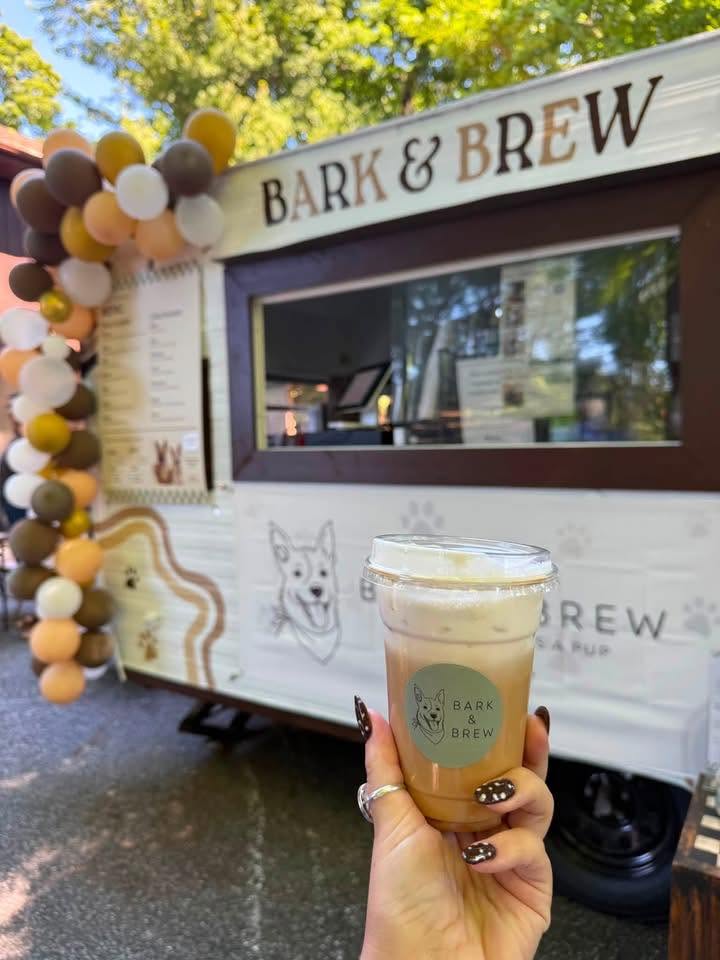 A hand holding an iced Bark & Brew coffee in front of the white trailer, decorated with a balloon garland and Bark & Brew signage